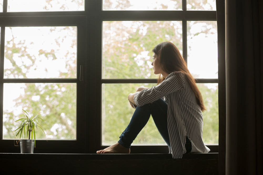 young girl, troubled by anxiety, sitting on sill embracing knees looking at window